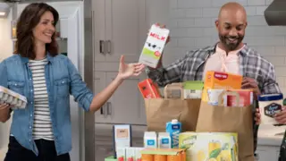 family unpacking their groceries. father is passing the mother a milk in paper carton to put in the fridge.